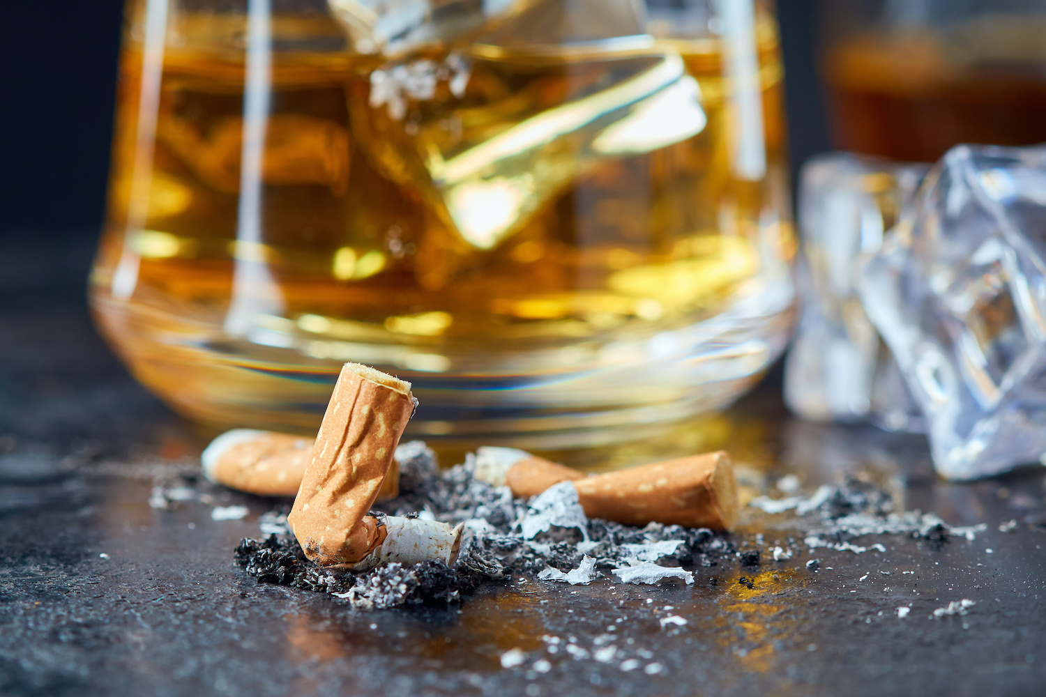 Cigarette butts crushed into ground, with a scotch glass full in the background