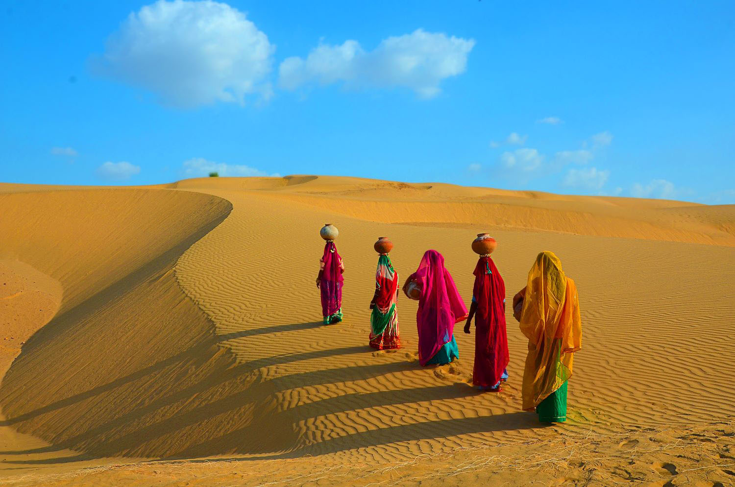 ndian women carrying heavy jugs of water on their head and walking on a yellow sand dune in the hot summer desert against blue sky