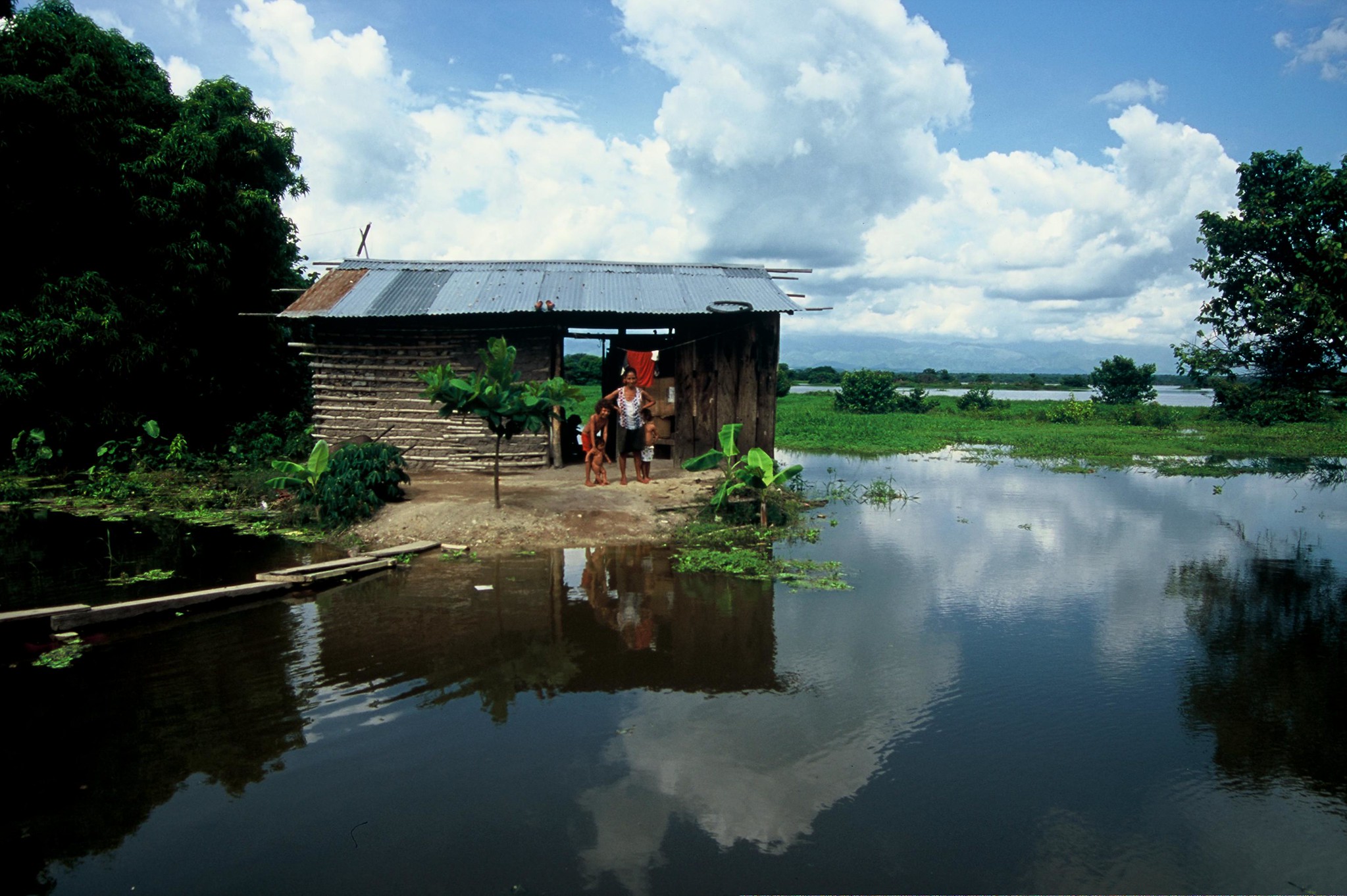 Mother and two young children standing outside of house surrounded by water in Colombia