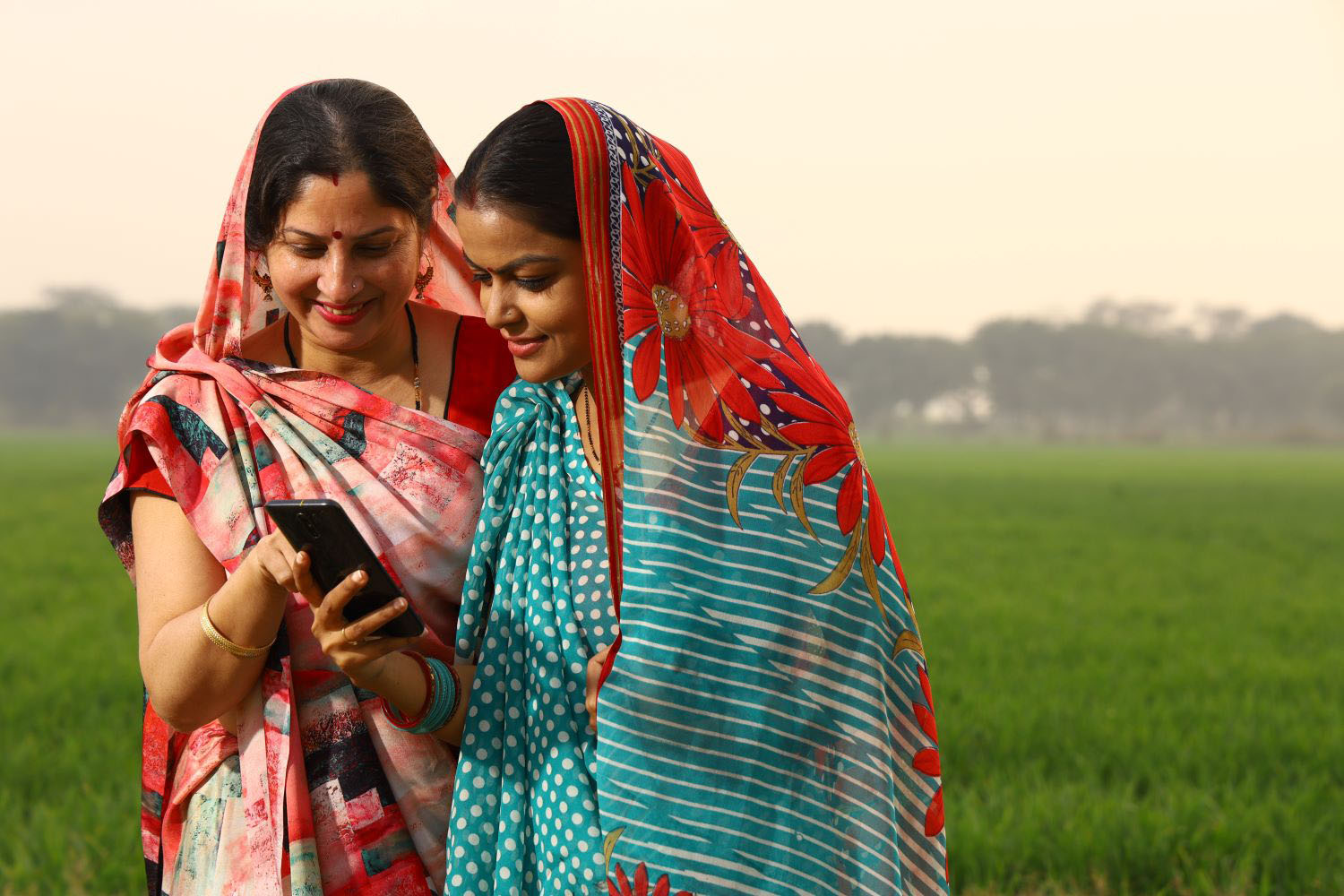 Happy rural Indian women standing in a mustard agricultural field and surfing through the mobile phone in their hand