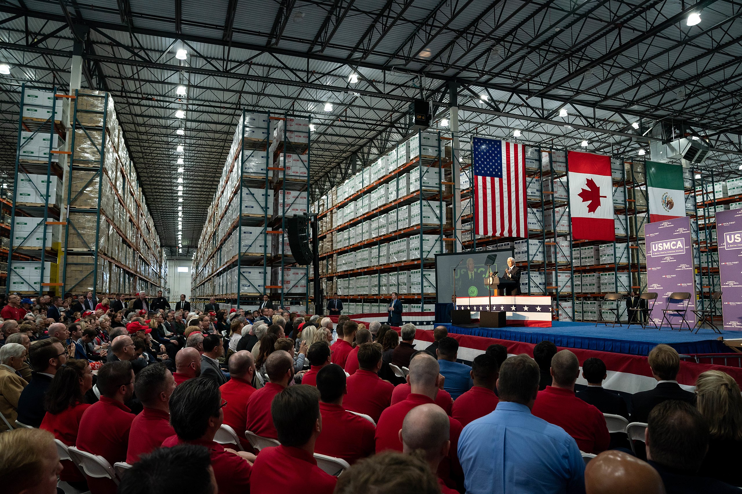 Politician speaks to full room at a factor with US Canada and Mexico flags in background