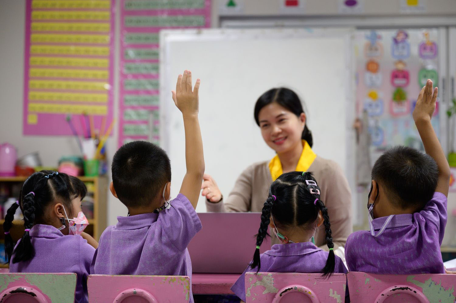 Kindergarten children raise their hands to answer questions in a classroom with a teacher