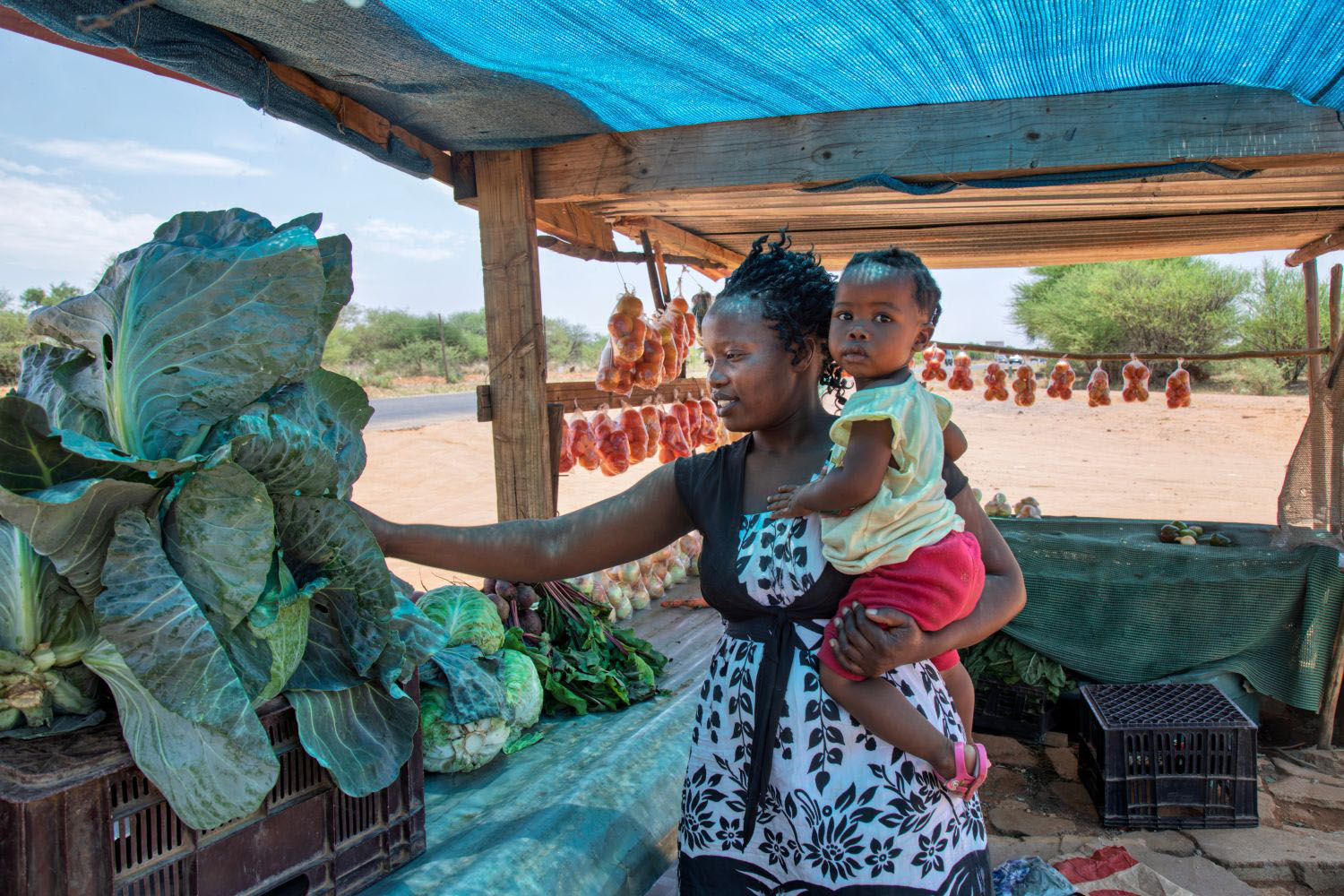 African woman vendor holding baby and selling vegetables from a wooden shack 