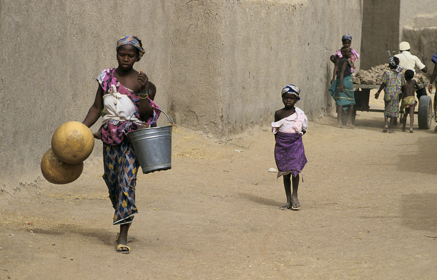 A woman carries water buckets while also holding an infant. Mali.