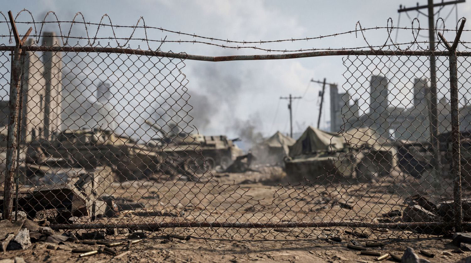 Gritty photograph looking through barbed wire at a hazy, smoke-filled war zone with military vehicles and tents