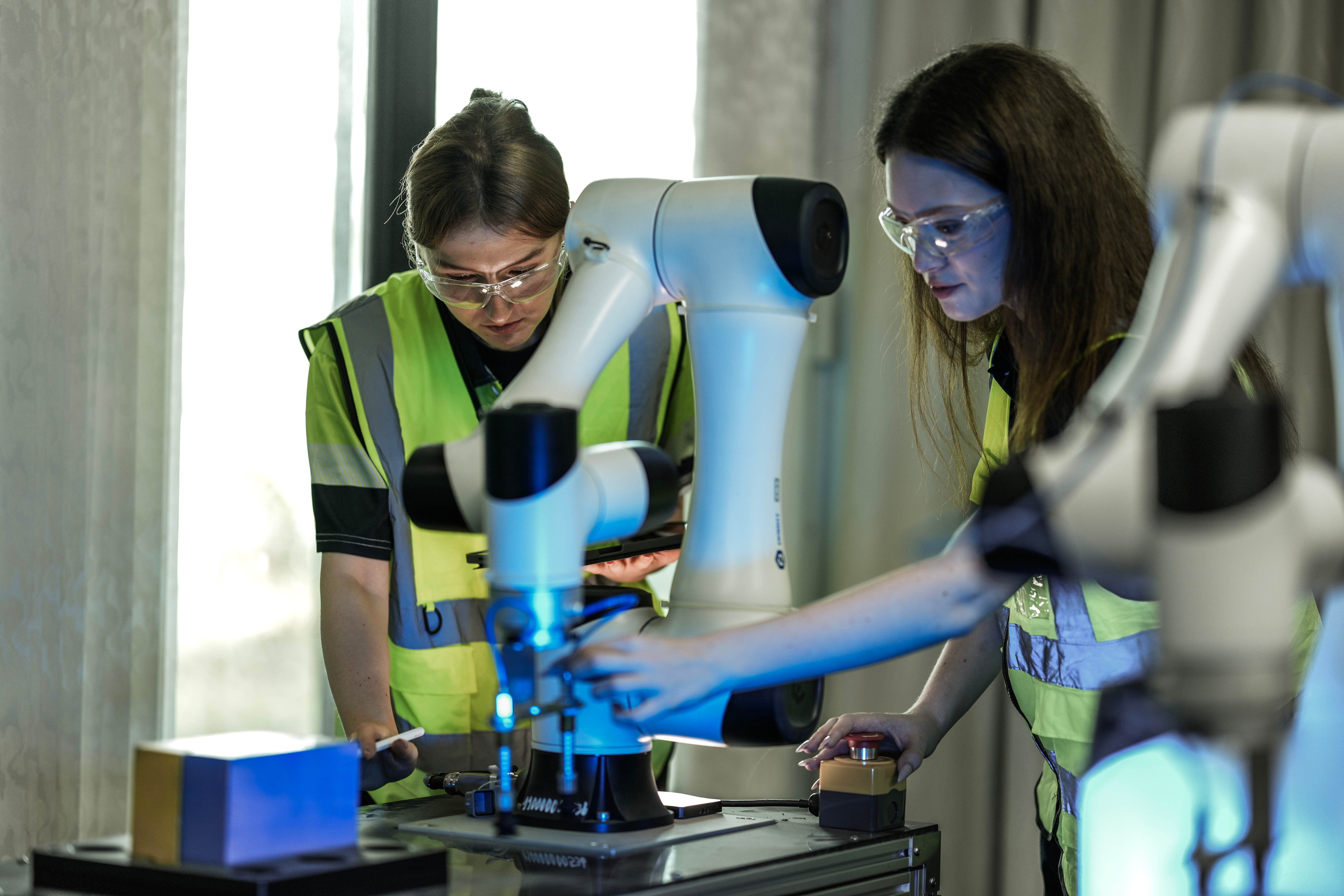 Two women engineers in technical clothing working in a lab with robotics