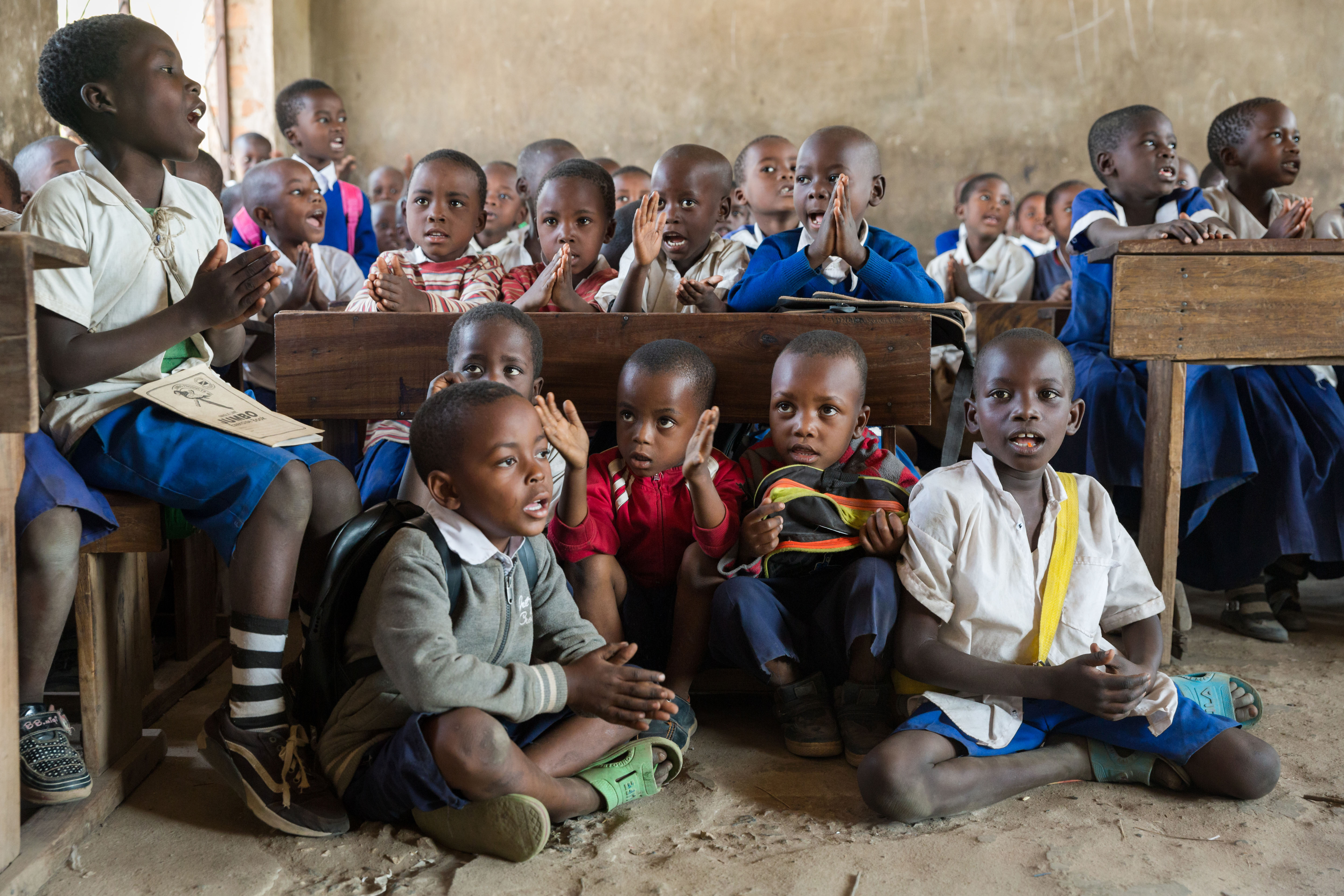 Children in a pre-primary classroom sit on the floor at the front of the classroom as the desks are full. 