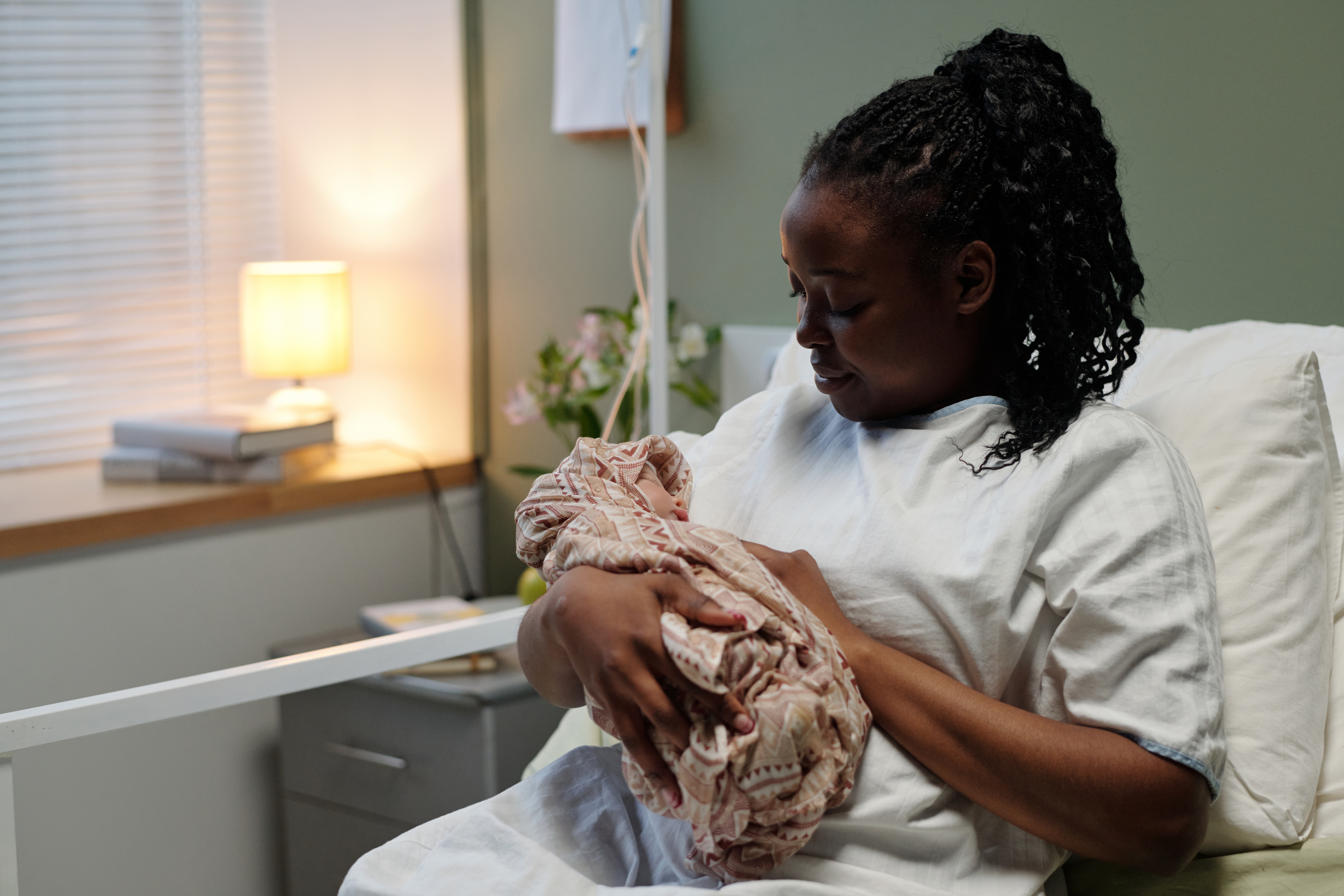 Black woman sitting in hospital bed in softly lit room, tenderly cradling her newborn while gazing at baby,