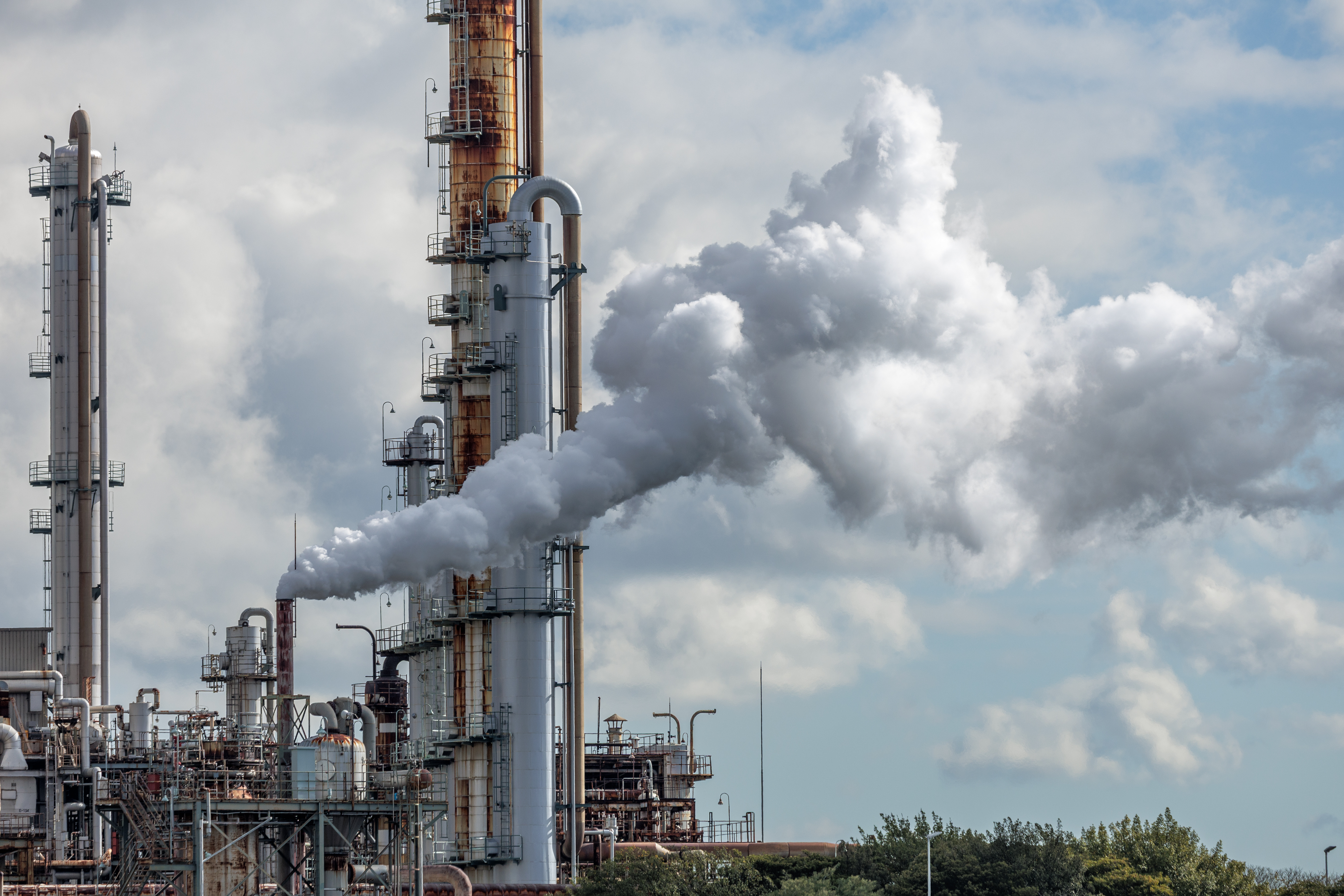 Industrial refinery plant complex emitting dense vapor cloud trail into partially overcast sky from rust-streaked refinery chimney