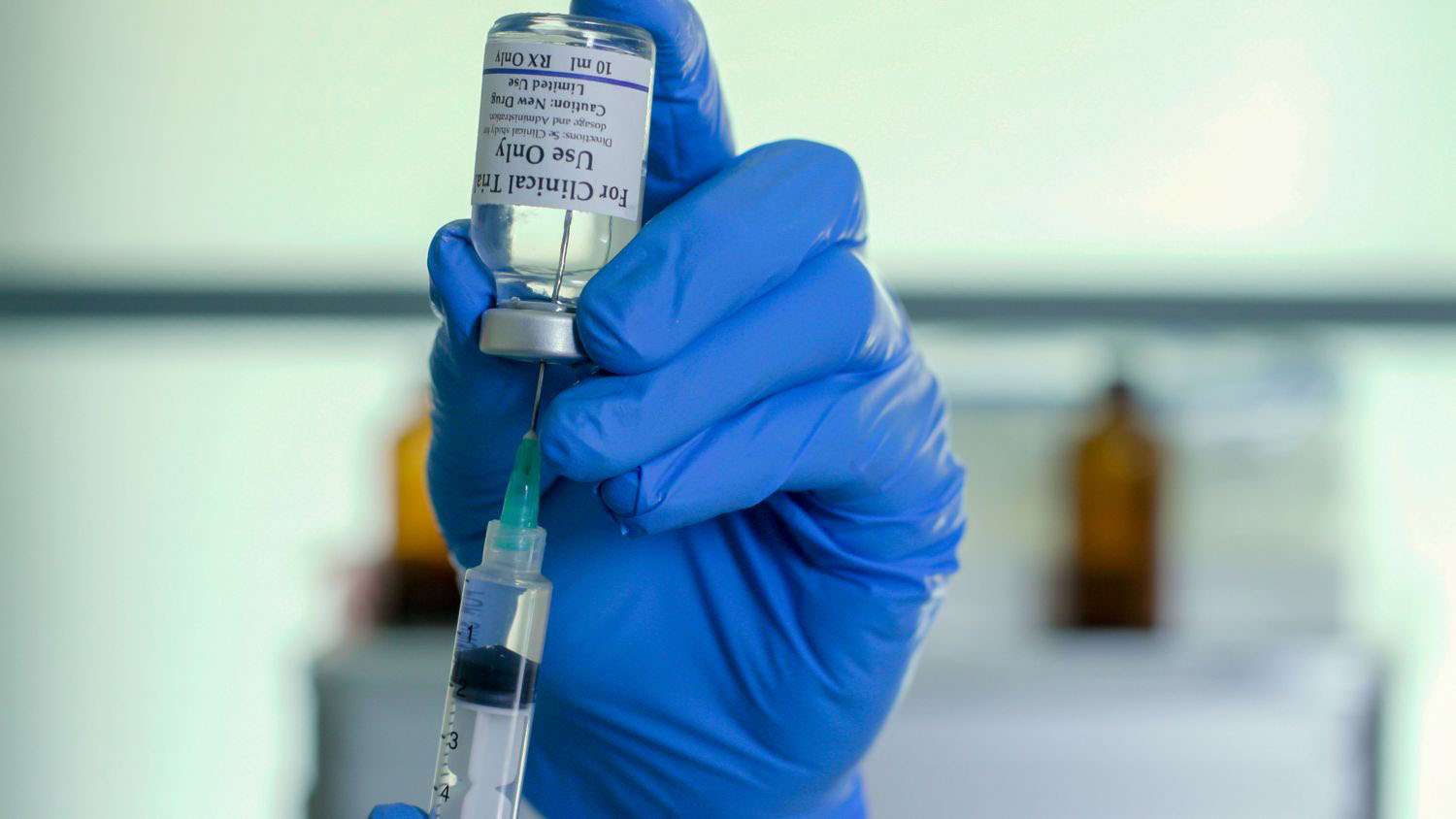 The gloved hand of a medical technician or researcher in search for a vaccine for COVID19 uses a syringe to extract from a vial labeled for clinical trial use.